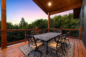 Terraza de madera con mesa y sillas, rodeada por una barandilla, con vista a un área boscosa.