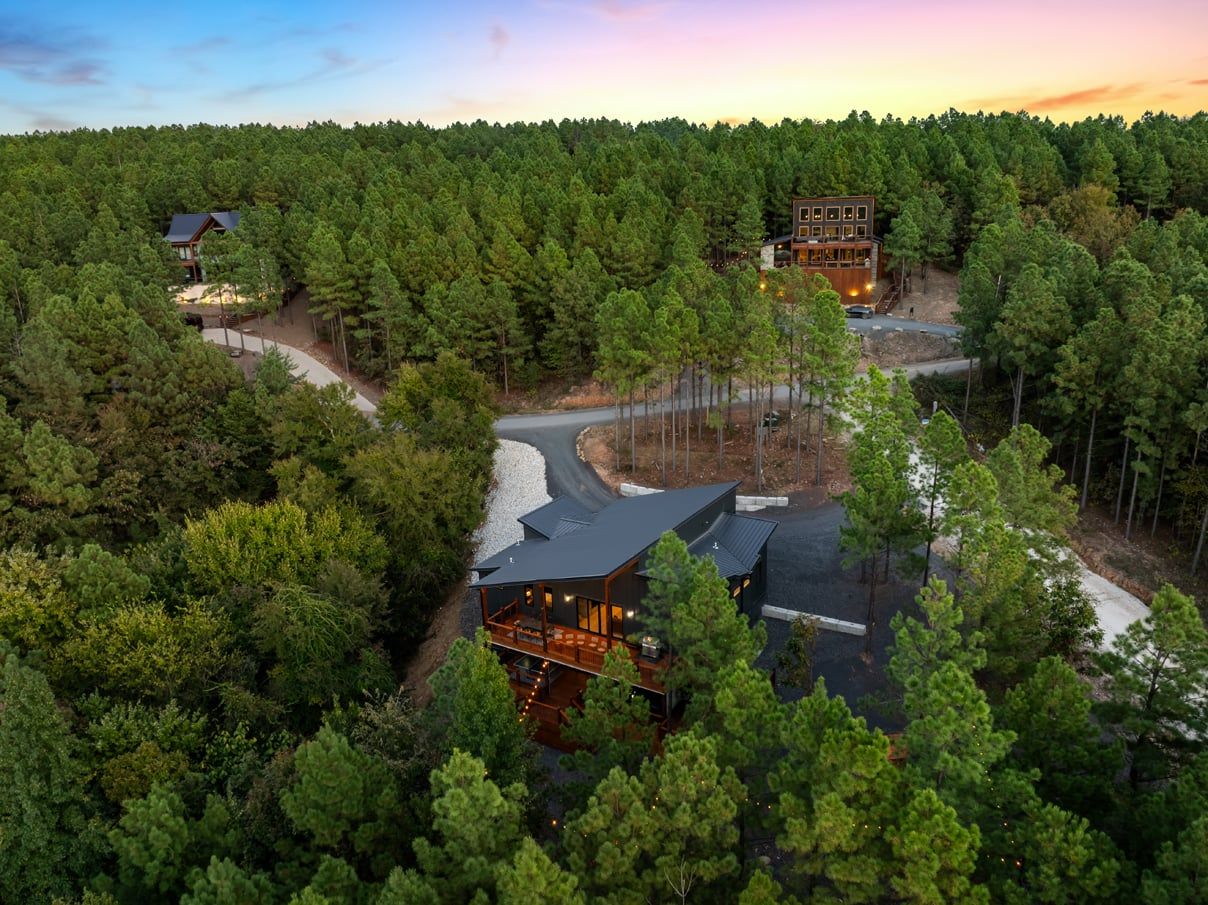 A modern lodge surrounded by dense forest, with a main building featuring a large deck and a smaller structure nearby.