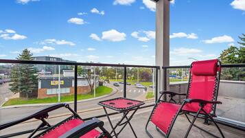 A balcony with two red chairs and a small table, overlooking a road with cars and buildings.