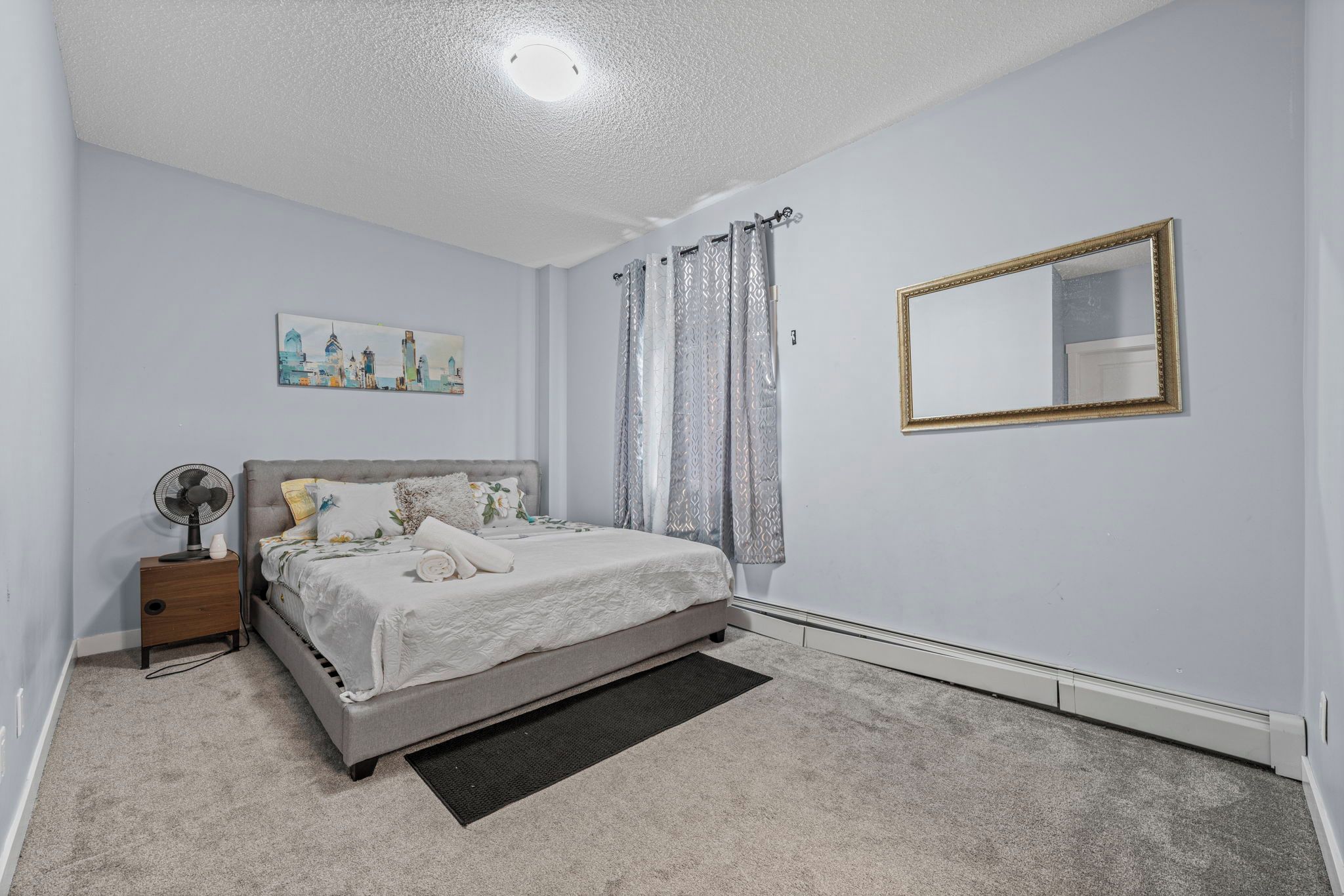 Une chambre à coucher avec un lit, une table de chevet, un ventilateur, un tableau, des rideaux et un miroir.