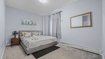 Une chambre à coucher avec un lit, une table de chevet, un ventilateur, un tableau, des rideaux et un miroir.