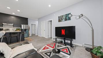 A modern living room with a flat-screen TV, a sofa, a coffee table, and a kitchen in the background.