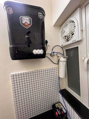 A black water heater with a vent, a white tiled wall, and a window with a view. - Private Apartment With Metro View on Highway (Mumbai)