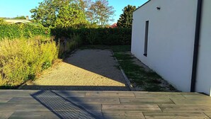 Ein gepflasterter Weg führt zu einem weißen Gebäude mit einem Fenster, umgeben von Grünflächen und blauem Himmel.