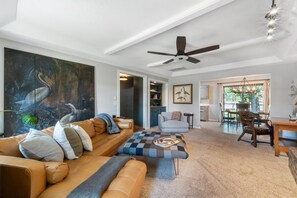 Sala de estar com uma grande pintura, ventilador de teto, sofá, mesa de centro e vista para a área de jantar.