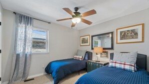 Une chambre avec deux lits, un ventilateur de plafond, une fenêtre avec des rideaux, une commode et des tableaux encadrés accrochés au mur.