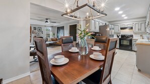 Un coin repas avec une table en bois dressée pour quatre personnes, entourée de chaises, d’un vase rempli de fleurs et d’un lustre moderne.