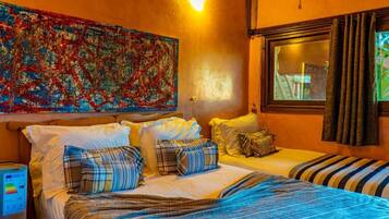 A hotel room with a bed, pillows, a blanket, a wall art piece, a window with curtains, and a bedside table with a lamp.