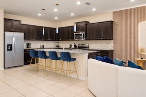 A modern kitchen with dark cabinets, a stainless steel refrigerator, a microwave, and a built-in oven. There are three blue bar stools and a white sofa with blue cushions. - 3029 Magical 10BR | Pool, Dino Slide & Games (Davenport)