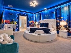 Un lit rond avec une tête de lit capitonnée, une table de chevet blanche et un fauteuil turquoise.