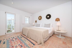 A bedroom with a bed, a bedside table with a lamp, a patterned rug, and a view through a window. - Les Terrasses B / Private Rooftop Pool in Tavira (Faro)