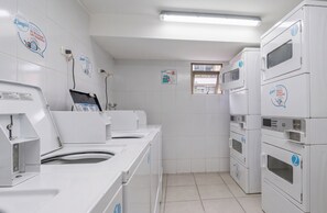 Un lavadero con varias lavadoras y secadoras, paredes de azulejos blancos y una ventana que deja entrar luz natural.