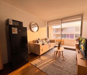 Un espacio habitable compacto con kitchenette, un sofá, una mesa de centro y un amplio ventanal con vista al exterior del edificio.