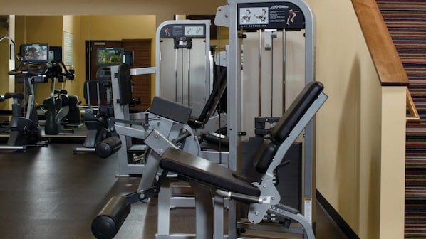 Uma academia com diversos equipamentos de exercício, incluindo uma máquina para extensão de pernas, e uma tela de televisão ao fundo.