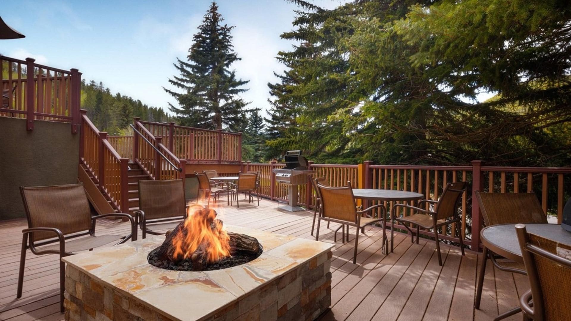 Una terraza exterior con un brasero de piedra, sillas y mesas de madera, rodeada por una barandilla de madera y árboles.