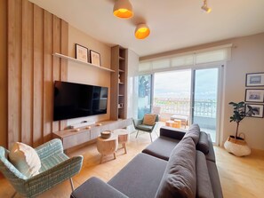 A modern living room with a flat-screen TV, a sofa, a chair, a small table, and a balcony with a view. - Coastal Paradise 2BR Marina View (Rio Hato)