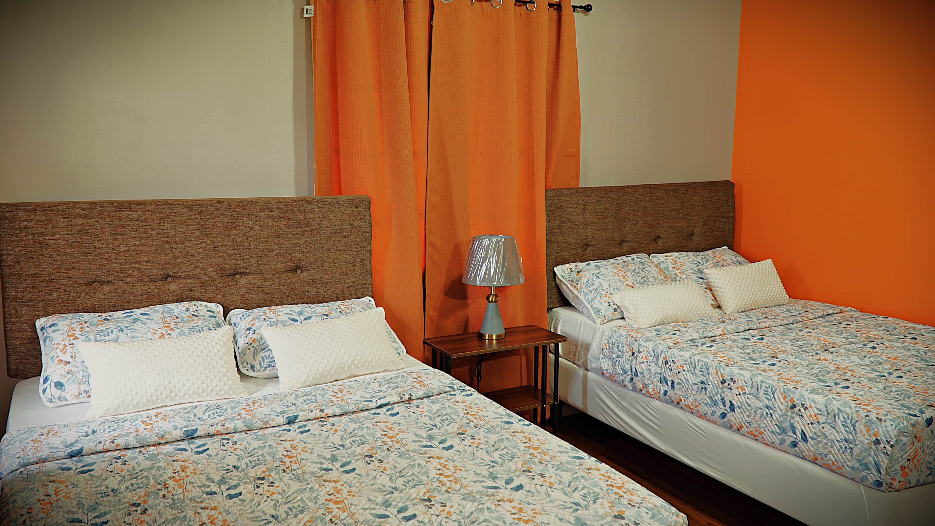 A hotel room with two beds, each with floral bedding, a nightstand with a lamp, and an orange curtain.