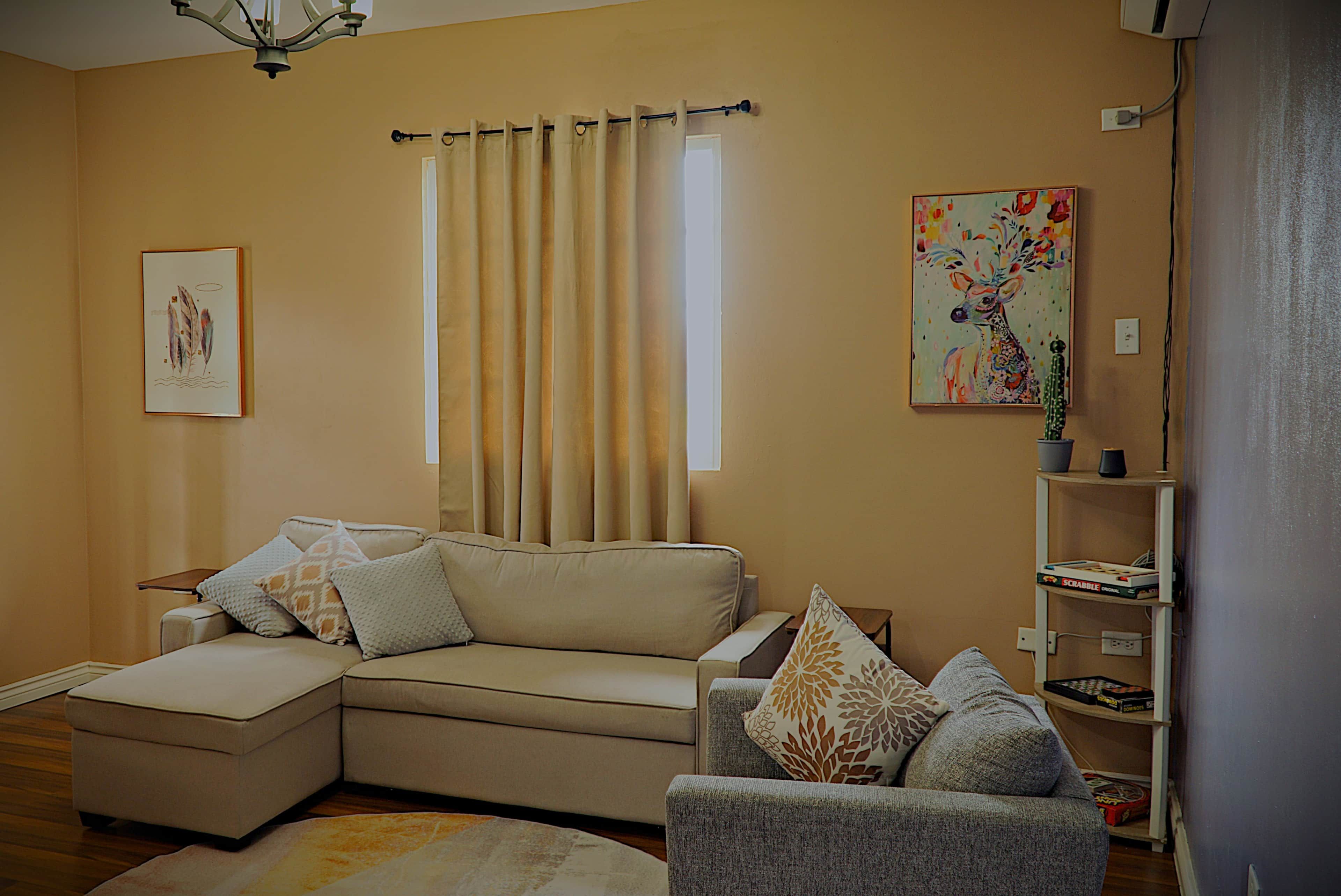 Ein Wohnzimmer mit einem beigefarbenen Sofa, einem grauen Sessel, einem gemusterten Teppich und einem Bild an der Wand.