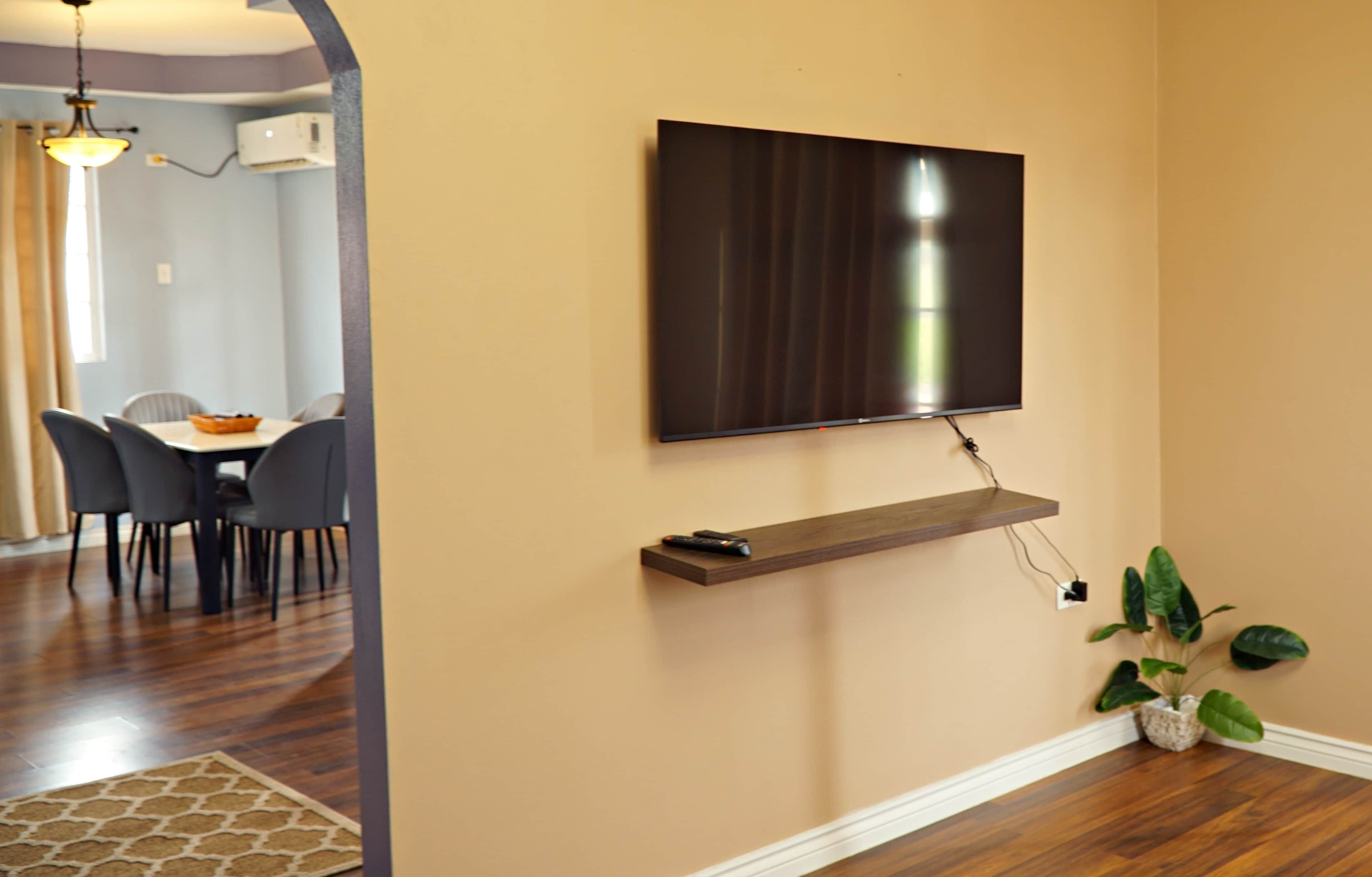 Ein modernes Wohnzimmer mit einem wandmontierten Flachbildfernseher, einem Holzregal darunter und einer Zimmerpflanze auf dem Boden.