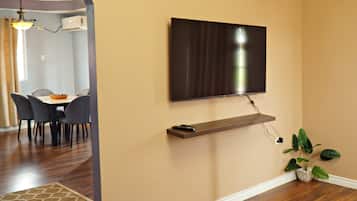 A modern living room with a flat-screen TV mounted on the wall, a wooden shelf below it, and a potted plant on the floor.