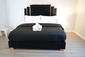 Una cama bien tendida con una colcha negra y almohadas blancas, una mesita de noche pequeña y una lámpara a la derecha.