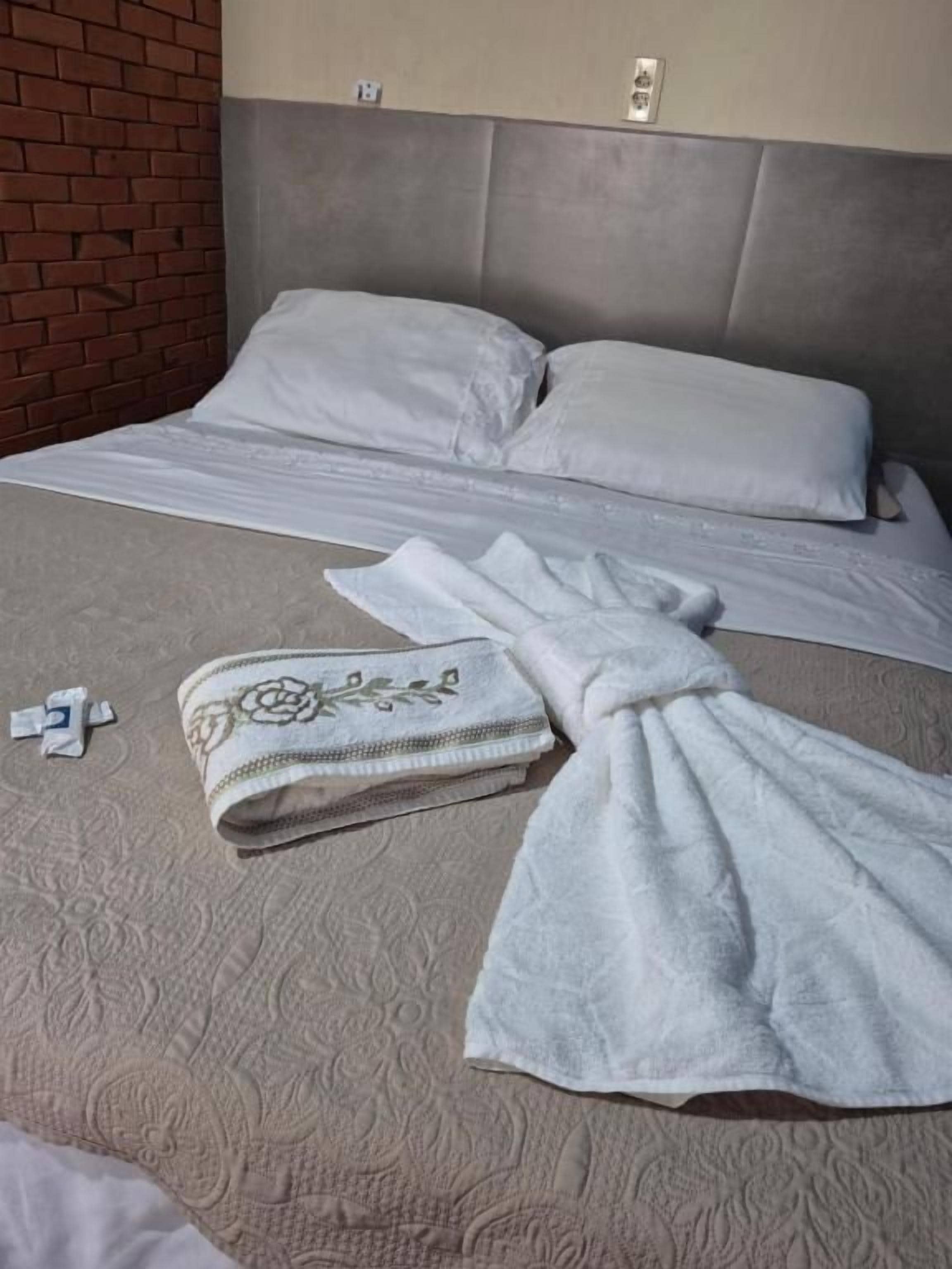 Uma cama com cabeceira cinzenta, manta branca e toalha dobrada no chão.