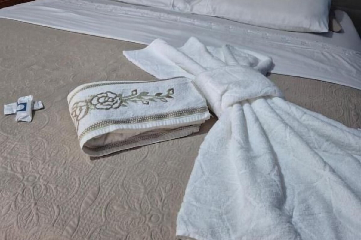 Una cama con cabecera gris, una colcha blanca y una toalla doblada en el suelo.