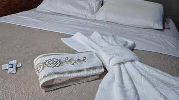 Uma cama com cabeceira cinzenta, manta branca e toalha dobrada no chão.