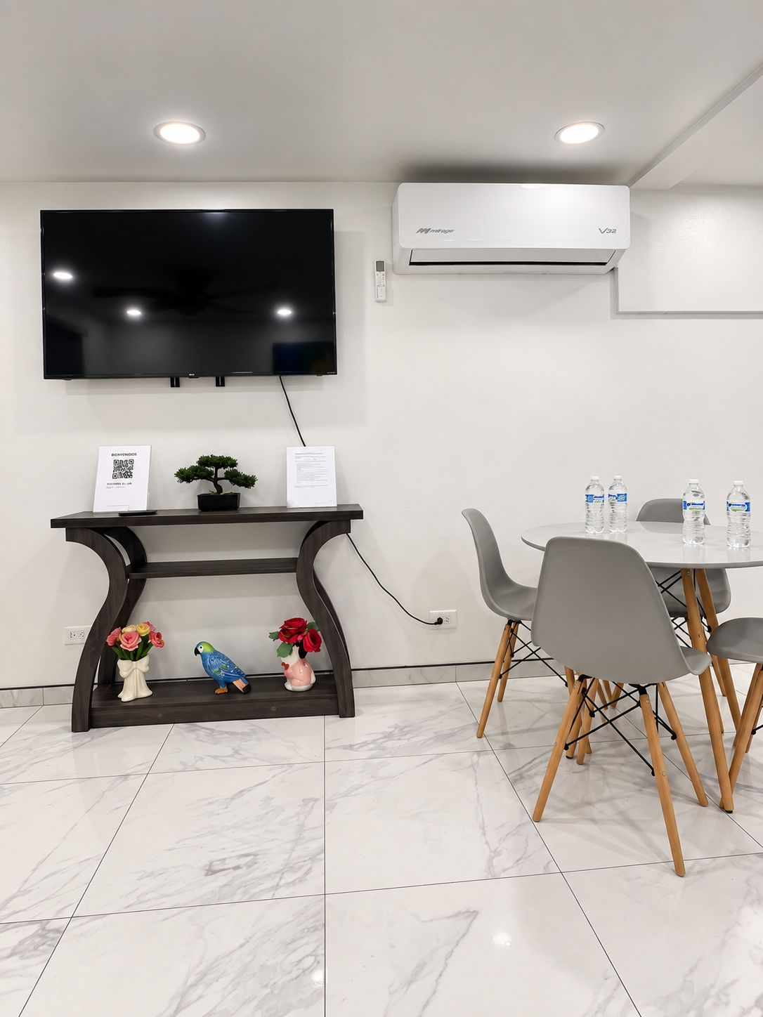 Quarto moderno com televisão de tela plana na parede, mesa consola de madeira escura com objetos decorativos, mesa de jantar redonda com cadeiras cinzas e um aparelho de ar condicionado.