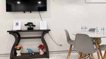 Quarto moderno com televisão de tela plana na parede, mesa consola de madeira escura com objetos decorativos, mesa de jantar redonda com cadeiras cinzas e um aparelho de ar condicionado.