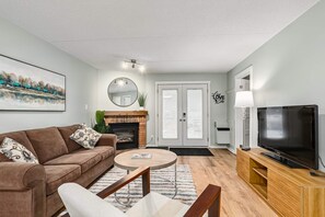 A living room with a brown sofa, a round coffee table, a TV, and a fireplace. - K's Condominium - Minutes to Ski, Trails, Shops, Beach (Collingwood)