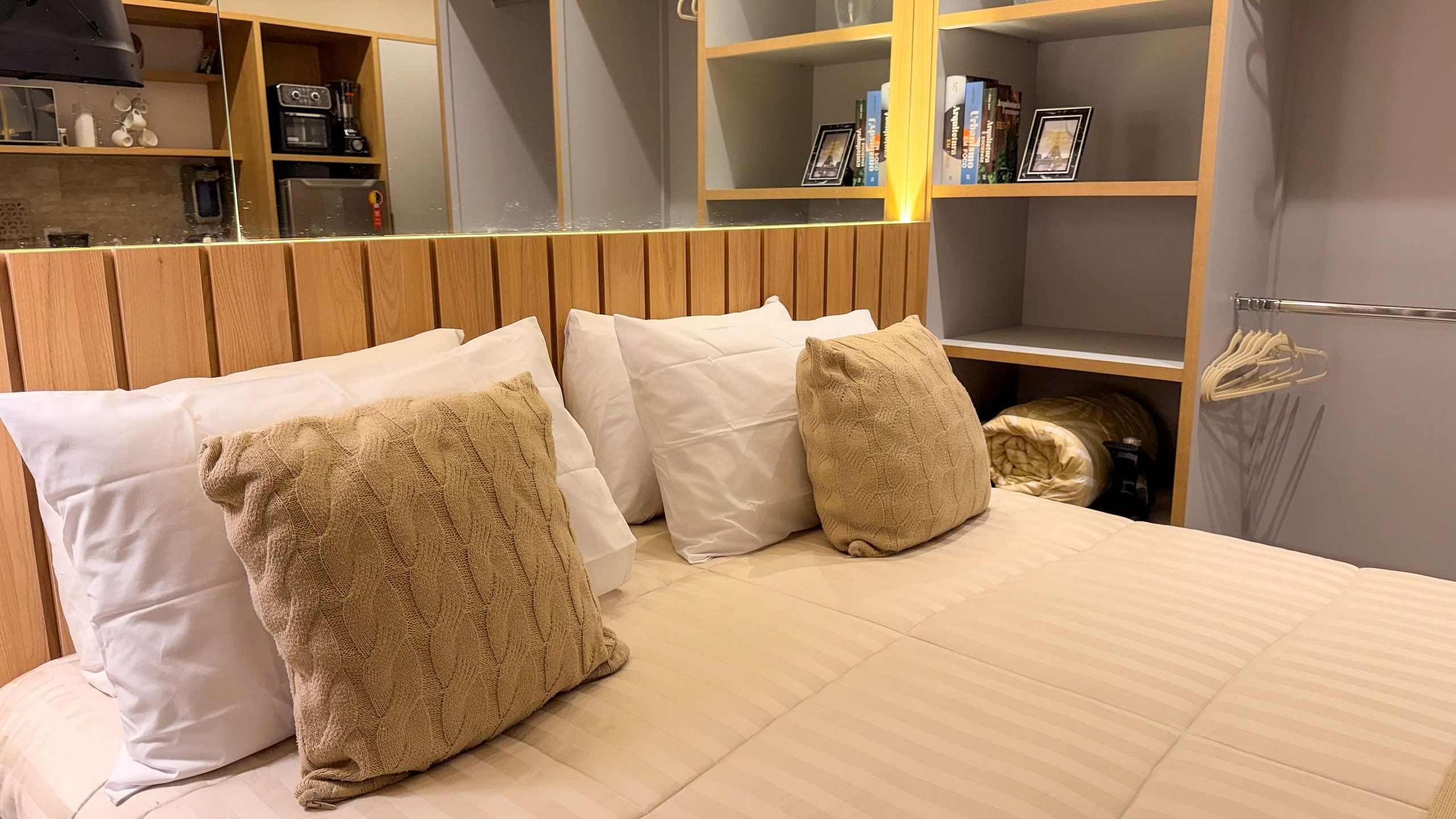 A hotel room with a bed, pillows, a wooden headboard, and a shelf with books and a picture frame.