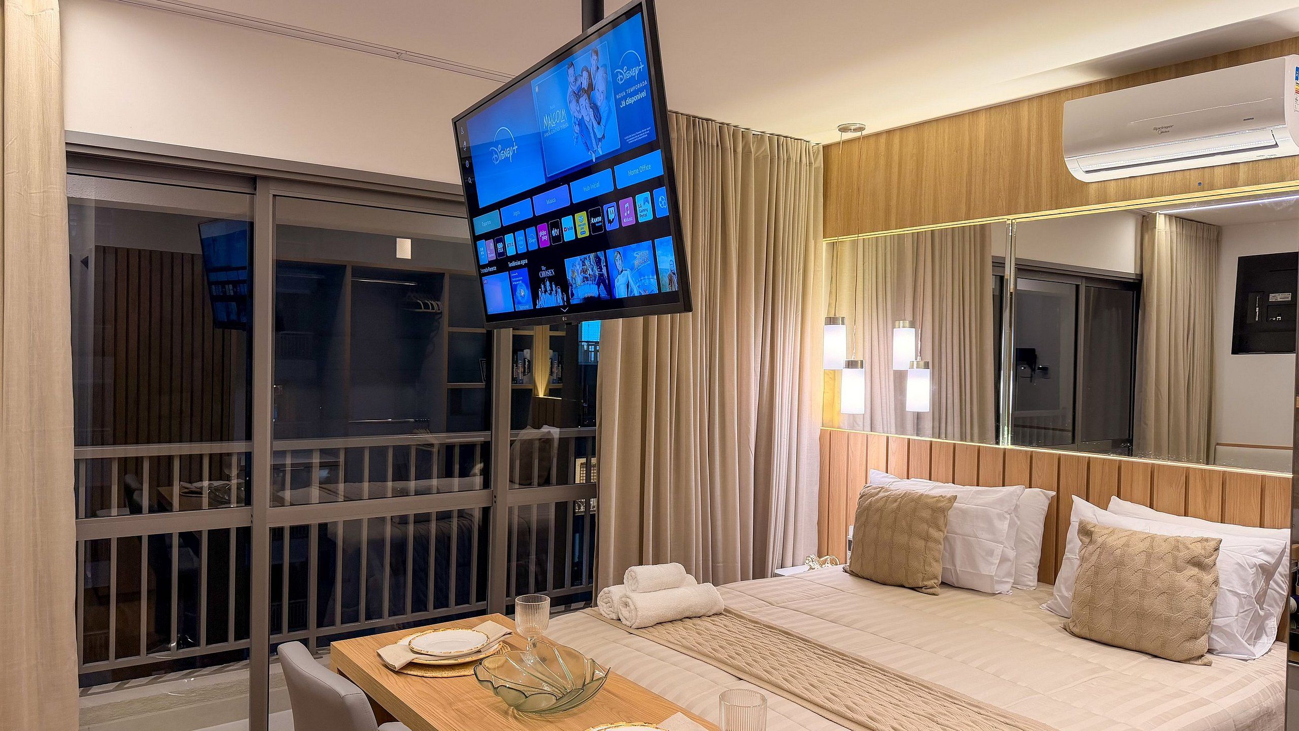 A hotel room with a bed, a TV mounted on the wall, a window with curtains, and a table with a glass.