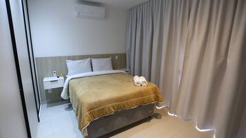 Quarto de hotel com cama, mesa de cabeceira, painel de controle e cortina.