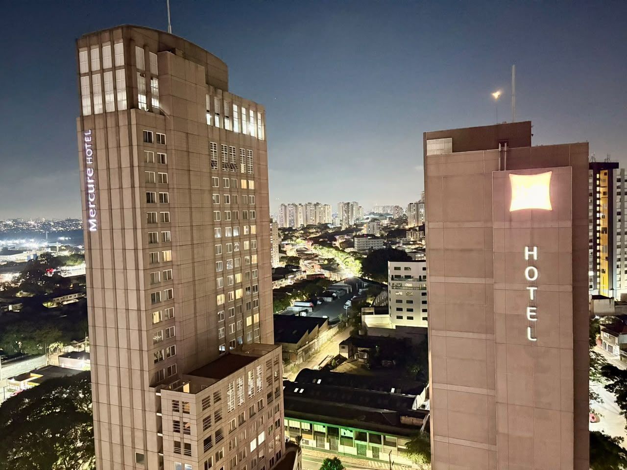A hotel building with a prominent 'HOTEL' sign, surrounded by high-rise buildings and a cityscape at dusk.