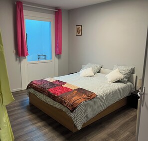 Une chambre à coucher avec un lit, une fenêtre avec des rideaux, une chaise placée à l’extérieur de la fenêtre et une petite table de chevet avec une lampe.