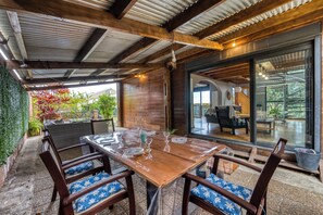 Uma área de refeições ao ar livre coberta, com uma mesa e cadeiras de madeira, cercada por paredes de vidro e vista para o interior.