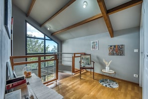 Ein geräumiges Zimmer mit großem Fenster, Holzbalken, einem Sessel, einem kleinen Tisch und Kunstwerken an der Wand.