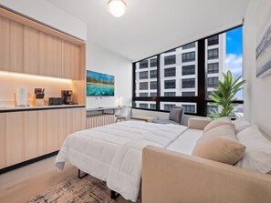 Une chambre moderne avec un grand lit, un bureau et une vue sur les immeubles à travers de grandes fenêtres.