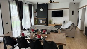 A modern living room with a dining area, a flat-screen TV mounted on the wall, and a ceiling fan.