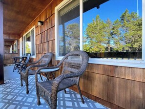 A covered outdoor seating area with wicker chairs and a table, wooden walls, and large windows overlooking trees. - Resting Beach Place | Luxurious Beachfront Condo (Neskowin)
