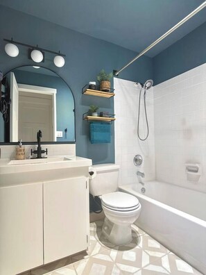 Une salle de bain avec des toilettes blanches, une baignoire et une douche. Il y a un miroir avec des lumières au-dessus, et une étagère avec des plantes.