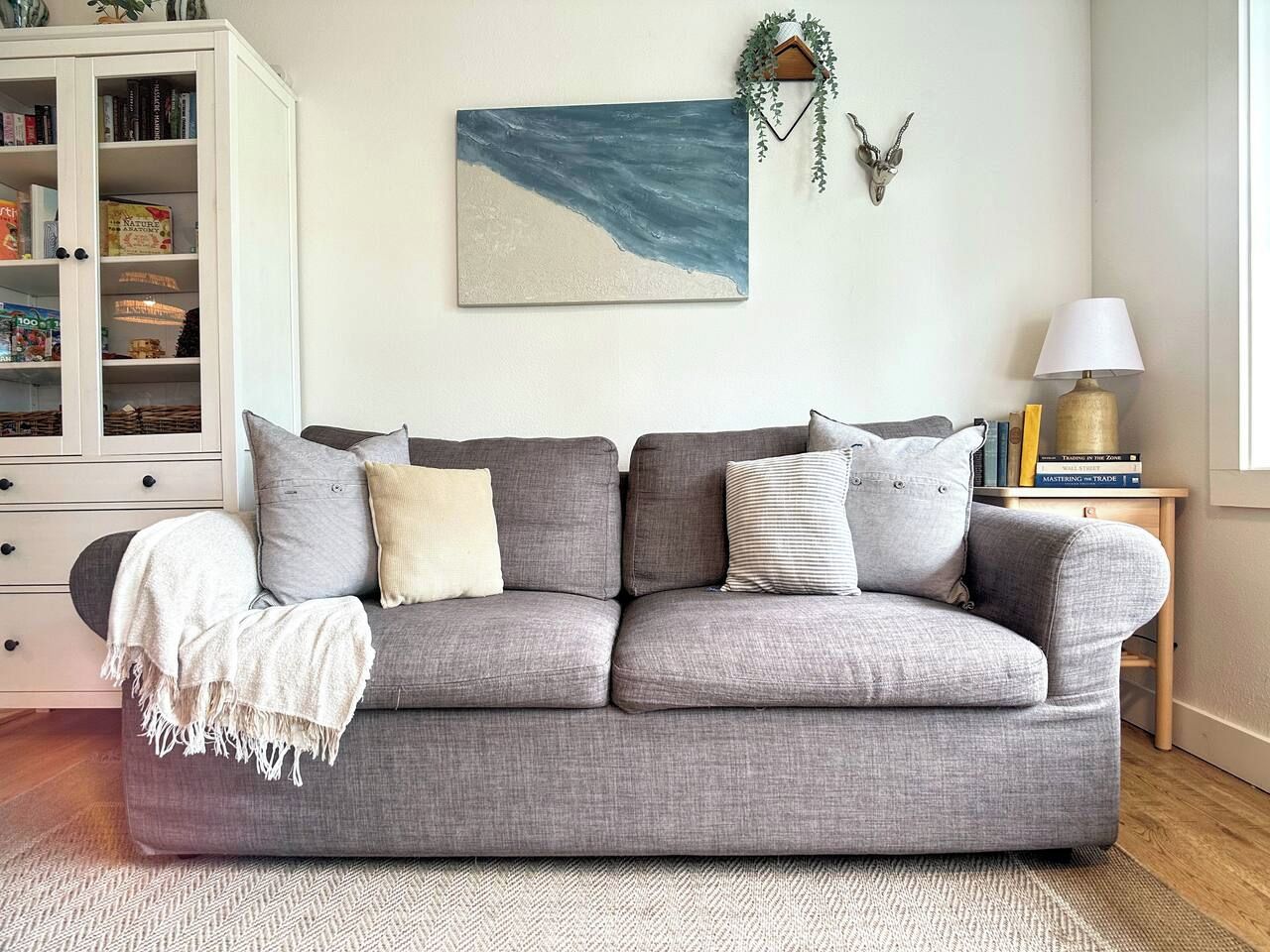 A grey sofa with multiple cushions, a white cabinet with drawers, a wall-mounted painting, a small table with books and a lamp, and a decorative plant.