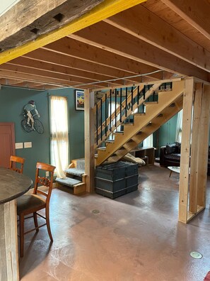 Ein geräumiges Loft mit freiliegenden Holzbalken, einer Treppe, einem Essbereich mit Tisch und Stühlen sowie einem Wohnzimmer mit einer Couch und einem Fahrrad.