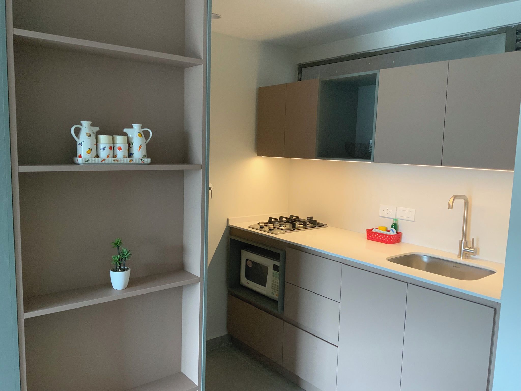 Eine kompakte Küche mit Gaskochfeld, Mikrowelle und Spüle. Oberhalb der Arbeitsplatte befinden sich Schränke und ein kleines Topfpflänzchen auf einem Regal.