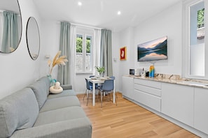 Ein modernes Wohnzimmer mit grauem Sofa, einem Esstisch mit Stühlen, einer Küchenzeile inklusive Mikrowelle und einem großen, an der Wand befestigten Fernseher.