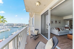A balcony with a view of a marina, a sliding glass door leading to a living area with a sofa and chairs, and a railing.