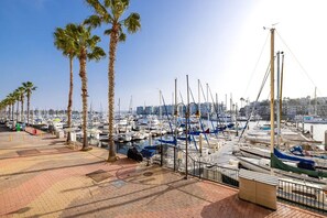 A marina with numerous boats docked, palm trees lining the walkway, and a clear view of the city skyline in the background.