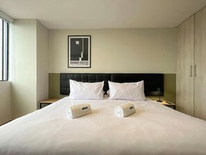 Ein Hotelzimmer mit einem großen Bett, zwei Kissen, einem gerahmten Bild an der Wand und zwei zusammengerollten Handtüchern auf dem Bett.