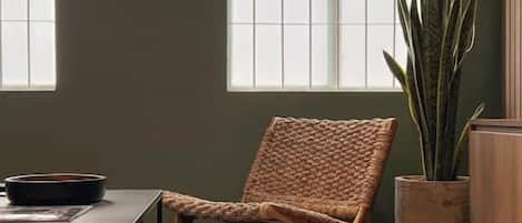 A room with a woven chair, a table with a bowl, a potted plant, and a framed picture on the wall.